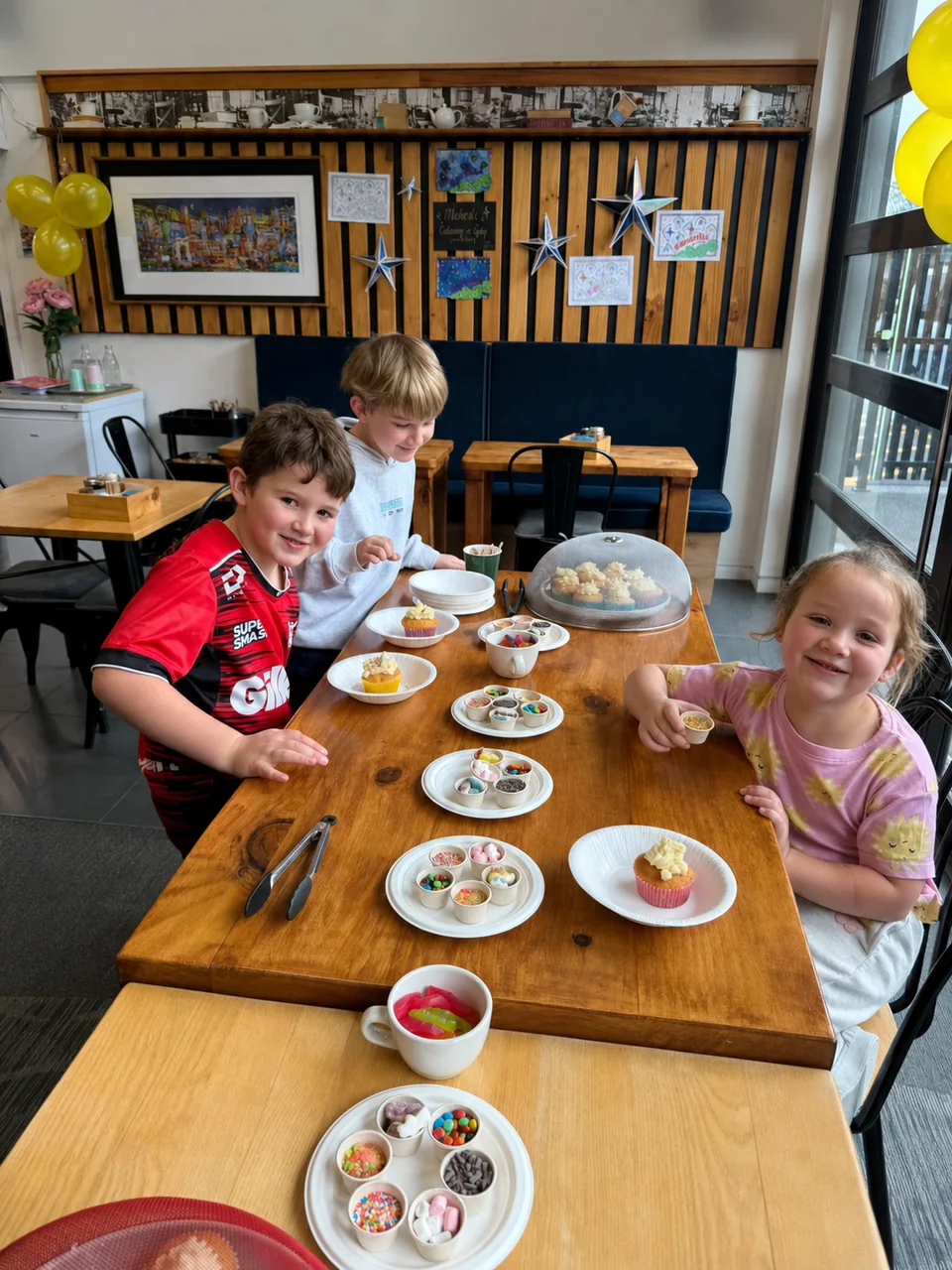 Children at a cafe table with cupcakes and bowls of toppings
